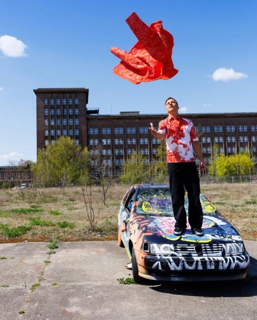 A man stands on a graffiti-covered car, tossing a red cloth into the air against a backdrop of a weathered building and blue sky.