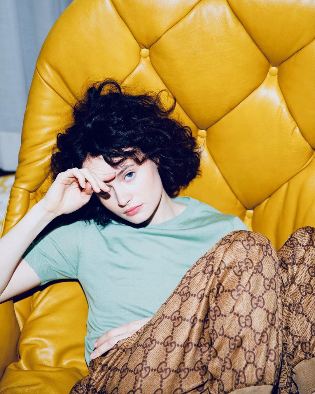 Woman with dark curly hair poses in a yellow armchair, wearing a mint shirt and patterned pants, looking directly at the camera.