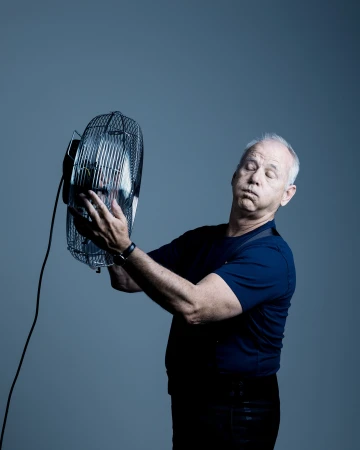 Older man holds a fan indoors, looking hot and bothered in a studio environment, with blue background and neutral lighting.