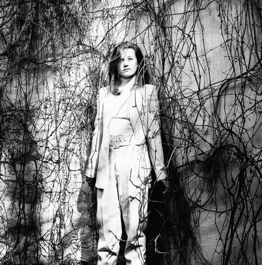 Monochrome portrait of a stylish adult woman in a suit, standing amidst climbing ivy, creating a stark, textured black and white image.