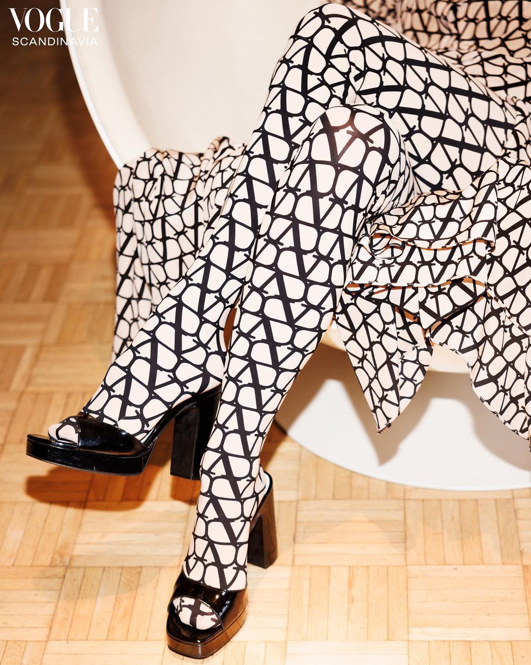 Legs of a person wearing patterned tights and platform heels while sitting in a modern chair on a wood floor with fashion logo.