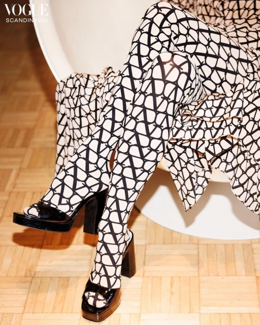 Legs of a person wearing patterned tights and platform heels while sitting in a modern chair on a wood floor with fashion logo.