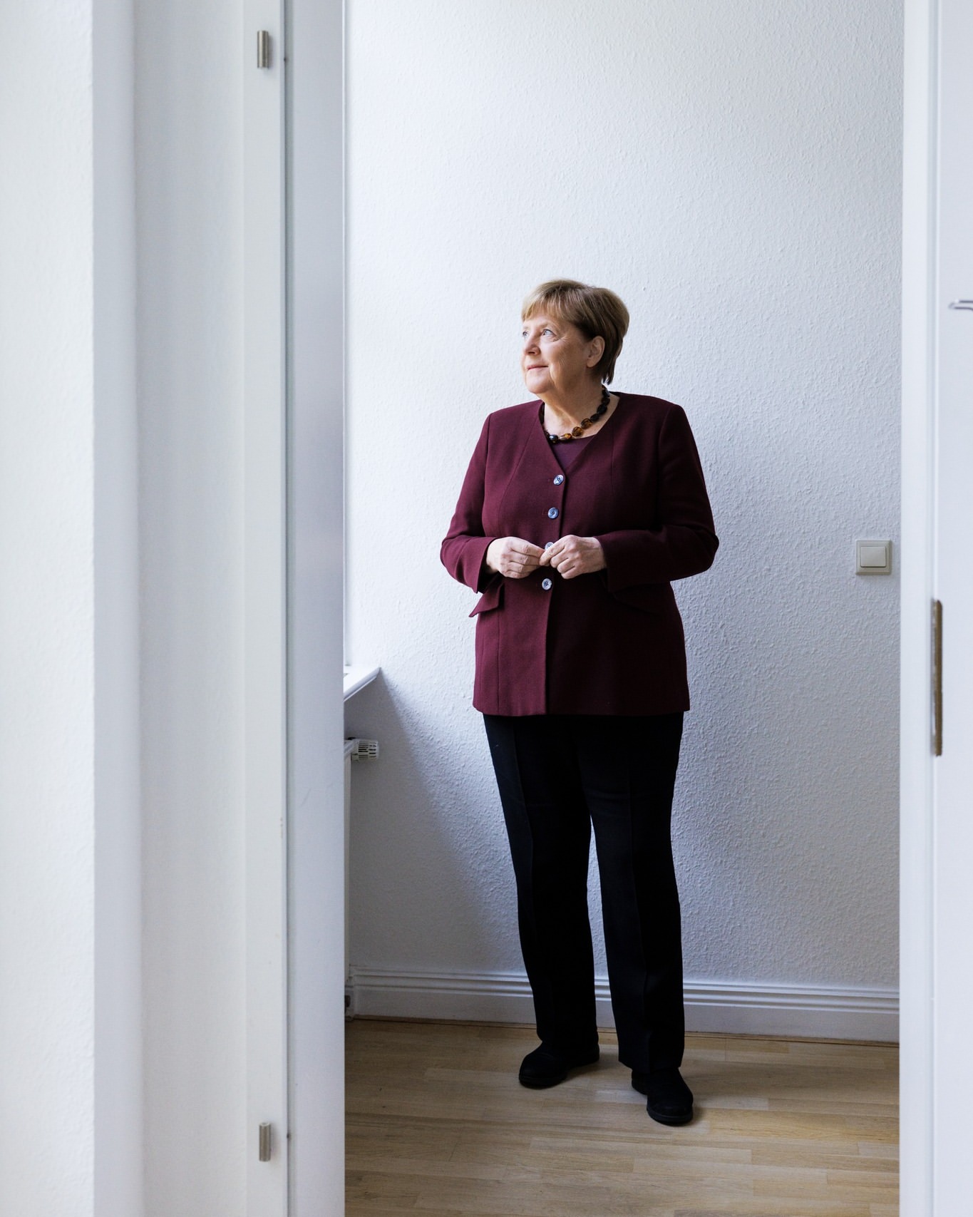 Indoor portrait of mature woman in burgundy jacket and black pants standing near doorway, looking out window in natural light.