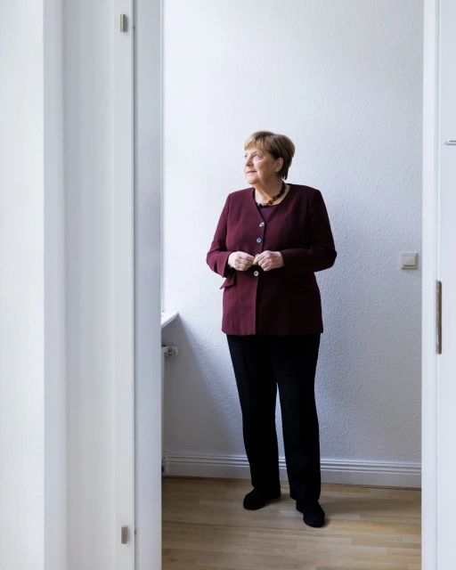 Indoor portrait of mature woman in burgundy jacket and black pants standing near doorway, looking out window in natural light.