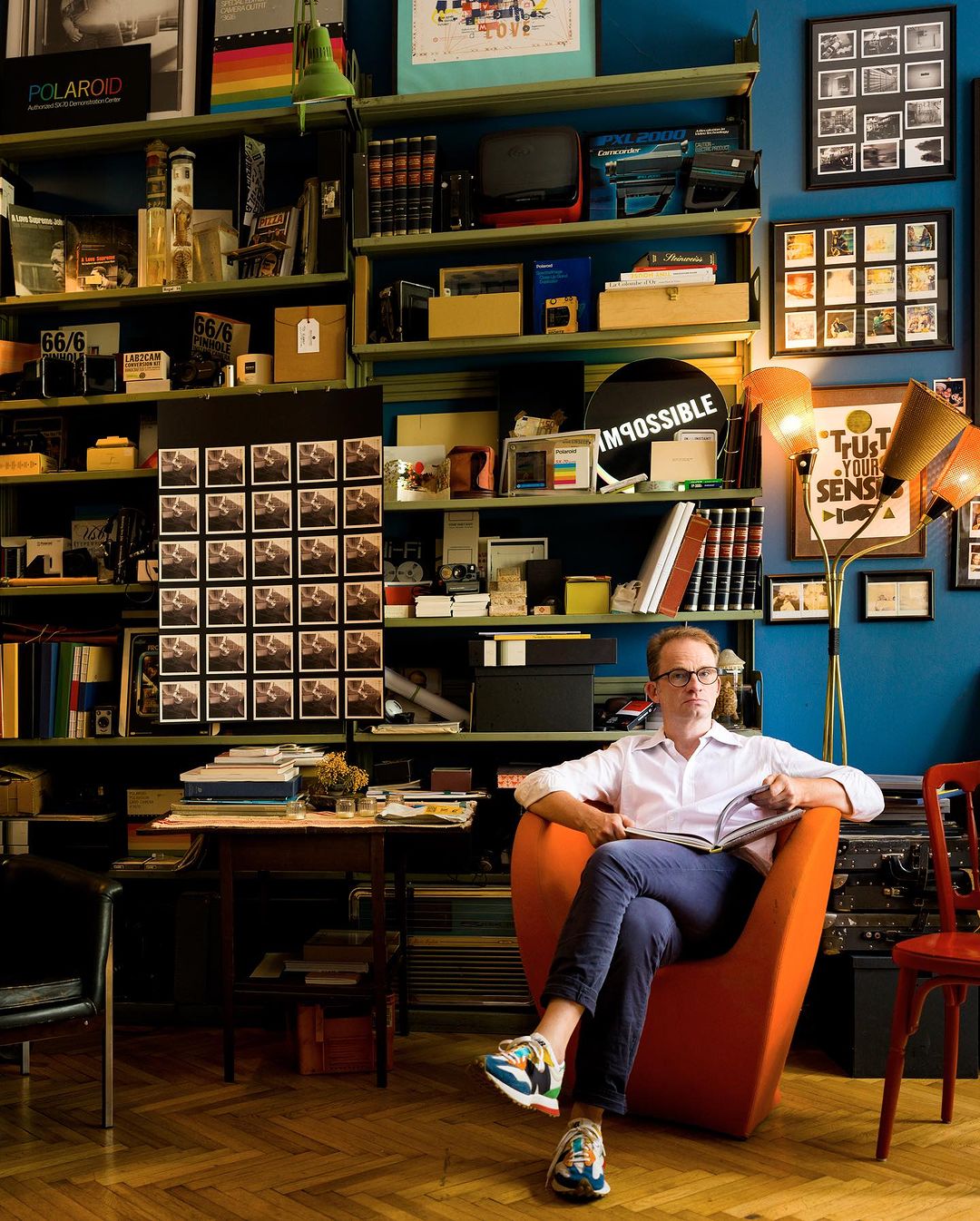 Adult male seated in an orange chair, holding a book, surrounded by shelves of books and objects in a brightly lit indoor space.