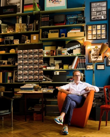 Adult male seated in an orange chair, holding a book, surrounded by shelves of books and objects in a brightly lit indoor space.