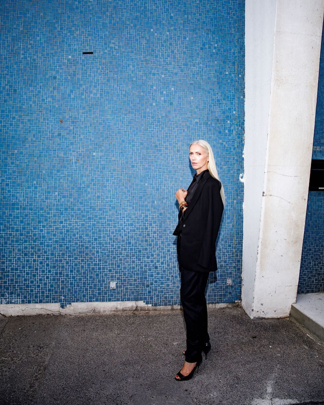 Adult woman with long blonde hair poses in a dark pantsuit against a blue tile wall outside, accessorized with jewelry and black heels.