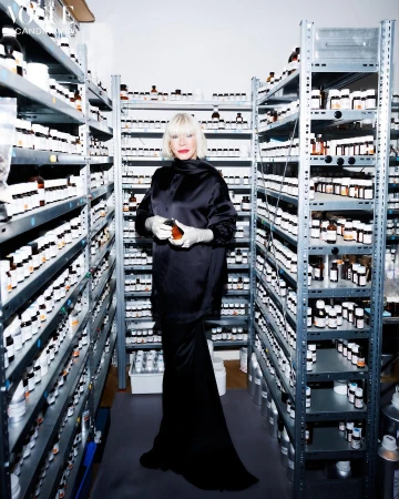 A fair-skinned adult woman with platinum blonde hair stands in a black gown in a brightly lit stockroom full of bottles on shelves.