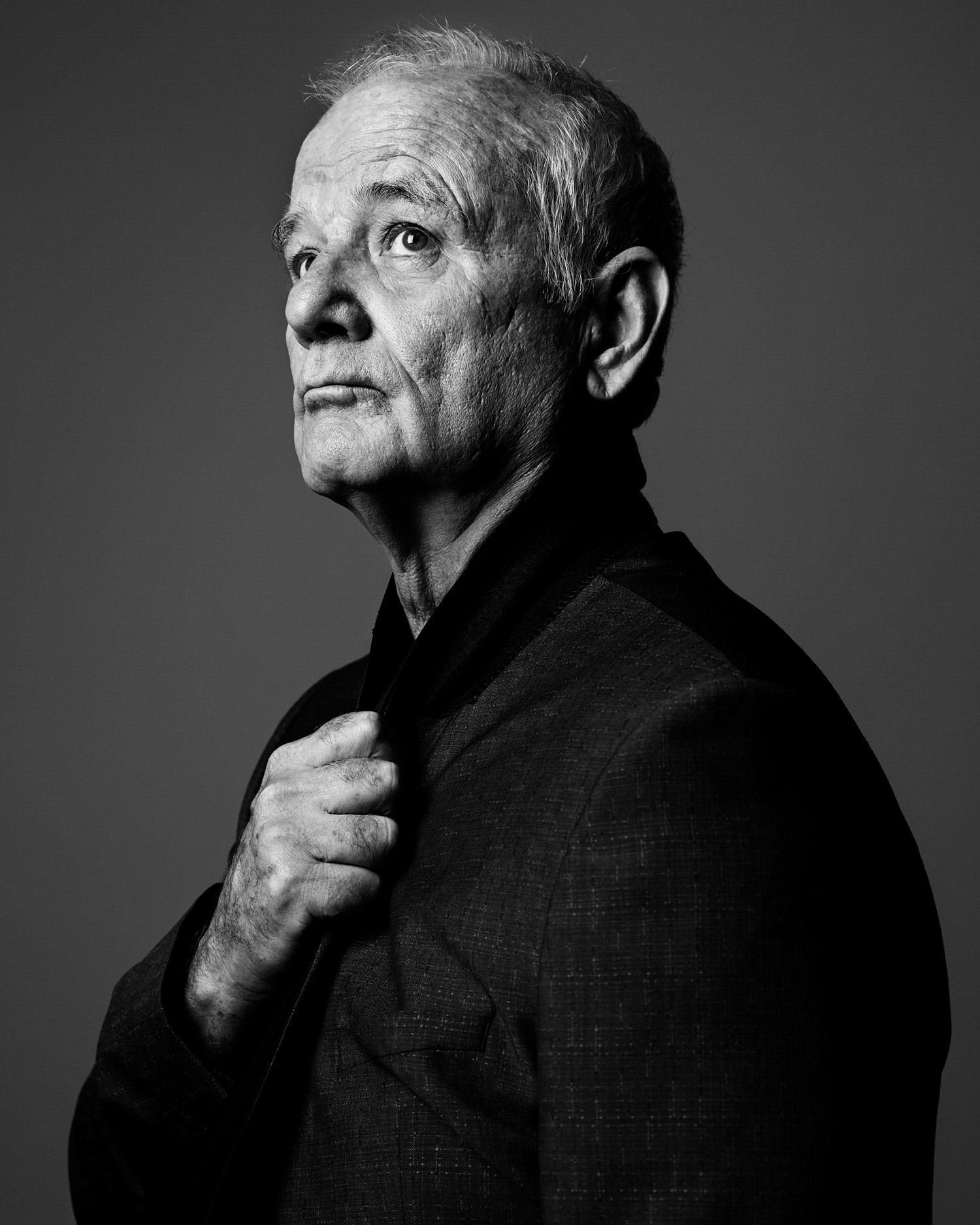 Monochrome portrait of an older gentleman with gray hair in a dark suit, looking upward with a hand on his necktie, against a gray backdrop.
