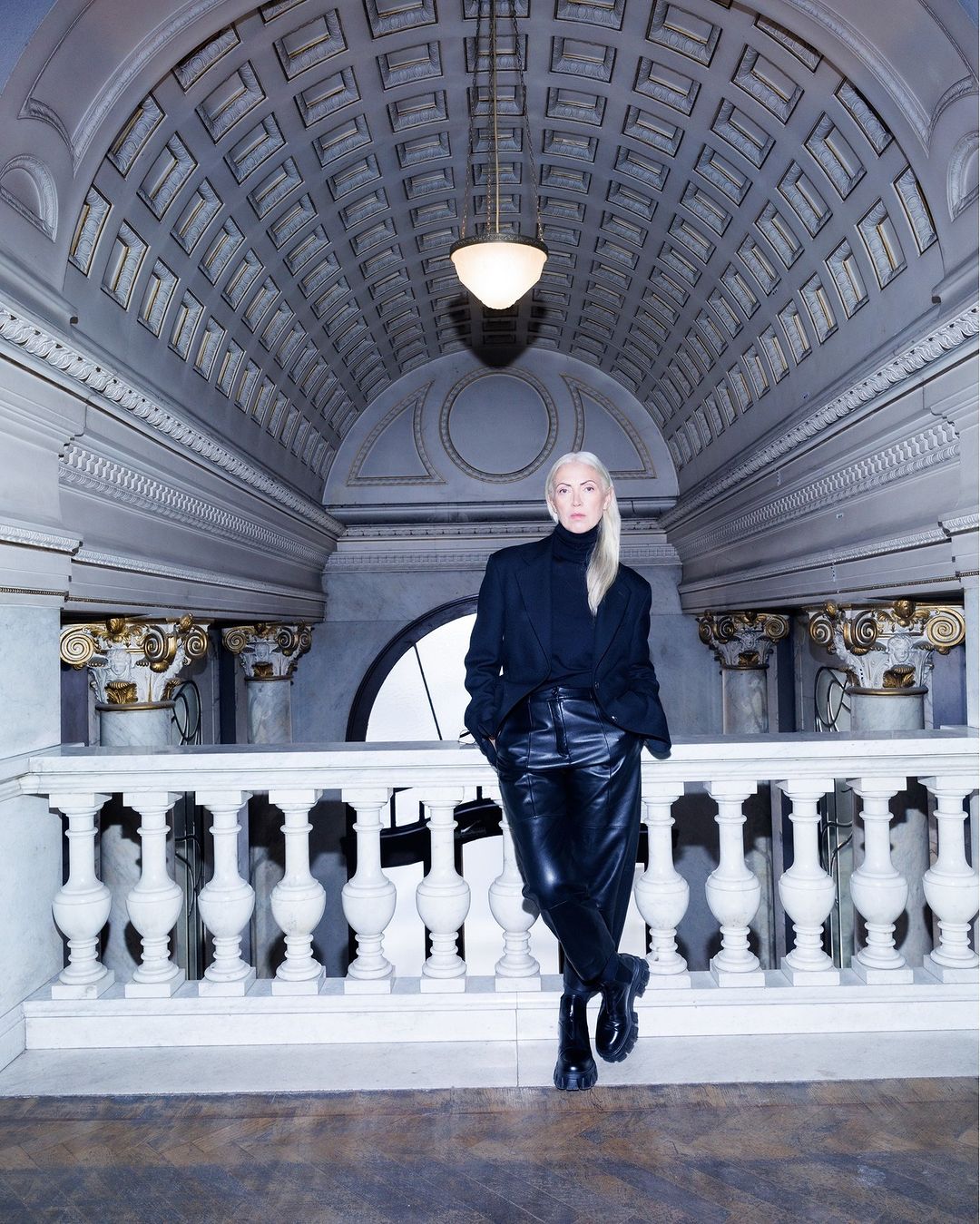 Adult female with blond hair poses in a black outfit indoors against a white stone railing and ornately designed ceiling with a lamp.