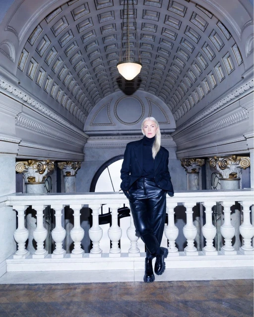 Adult female with blond hair poses in a black outfit indoors against a white stone railing and ornately designed ceiling with a lamp.