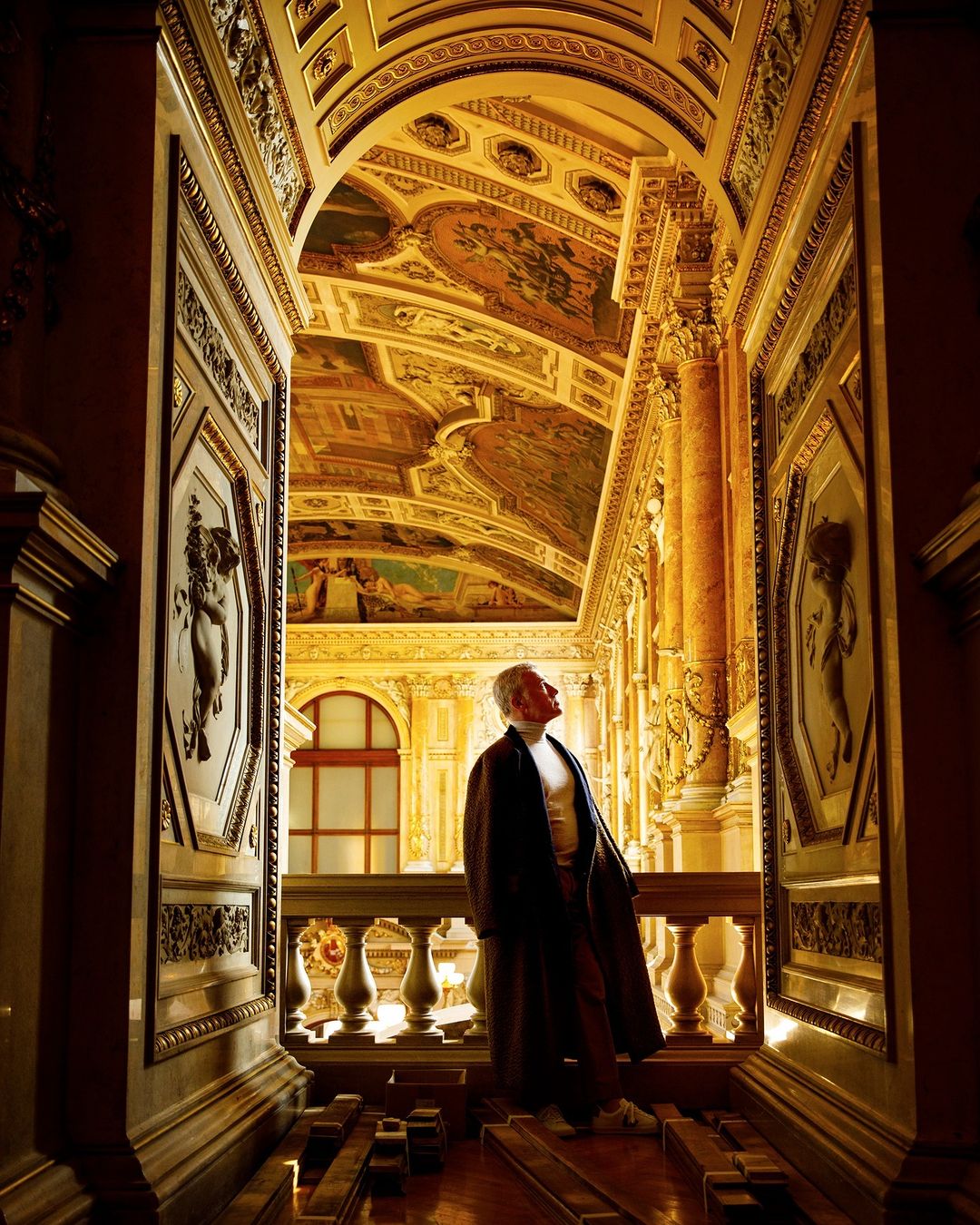 Mature adult in a long coat looking at ornate ceiling in a golden hall. Architecture and artistic details create a dramatic scene.