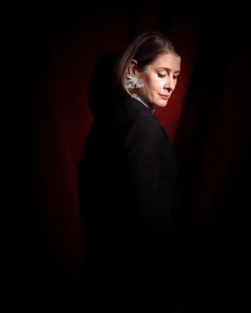 Portrait of a woman with blonde hair, wearing decorative earrings and a black top, posing in front of a dark red background.