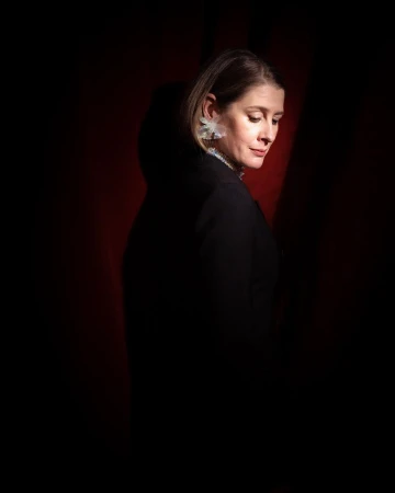Portrait of a woman with blonde hair, wearing decorative earrings and a black top, posing in front of a dark red background.