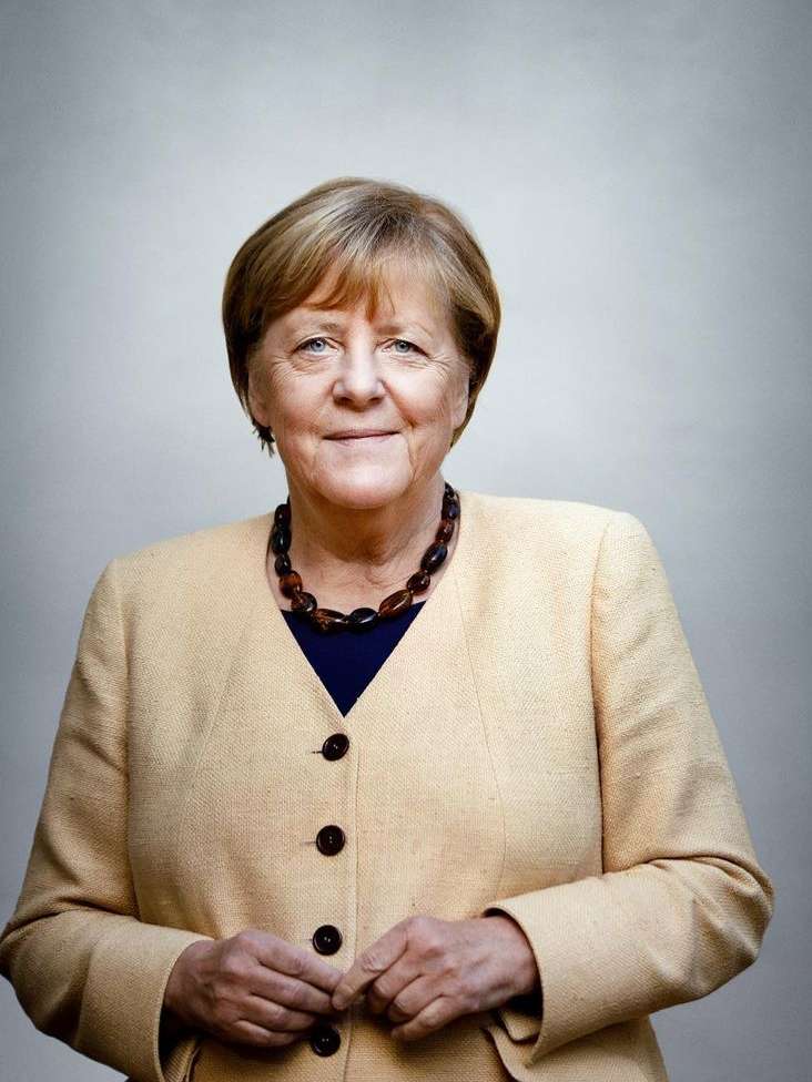 Headshot of an adult woman with short blonde hair, wearing a tan jacket, a dark top, and a brown bead necklace in front of a plain backdrop.