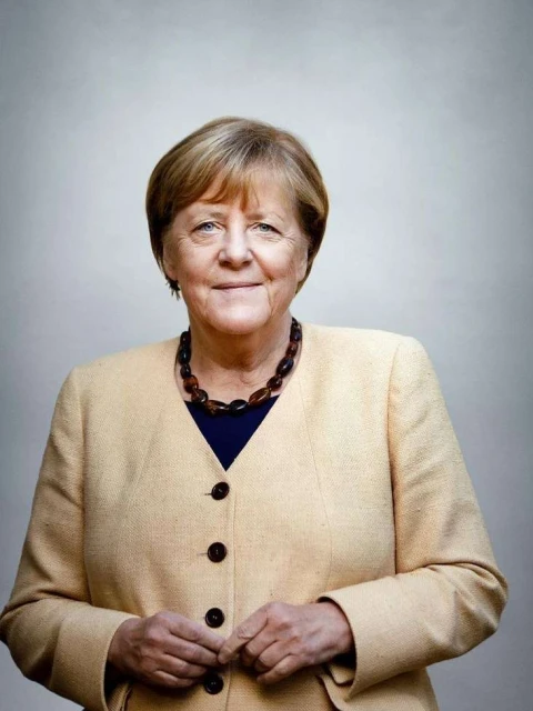Headshot of an adult woman with short blonde hair, wearing a tan jacket, a dark top, and a brown bead necklace in front of a plain backdrop.
