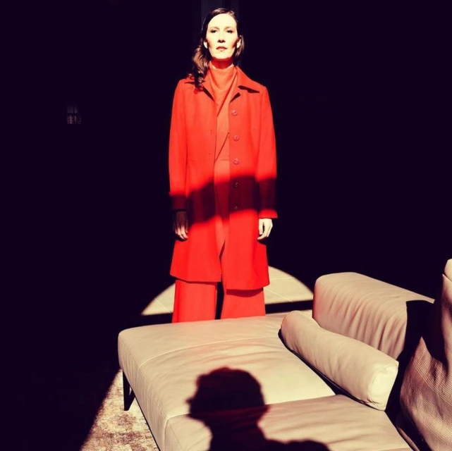 Adult female in red coat and pants stands in a room with a beige couch illuminated by direct sunlight in a high-contrast, editorial fashion shot.