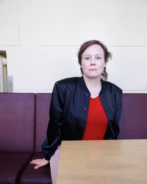 Indoor portrait of a woman in a black jacket and red shirt sitting at a light-colored wooden table with a mauve leather booth.