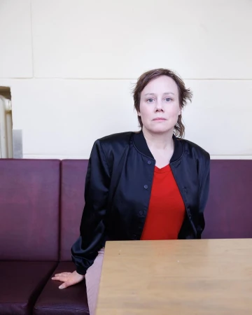 Indoor portrait of a woman in a black jacket and red shirt sitting at a light-colored wooden table with a mauve leather booth.
