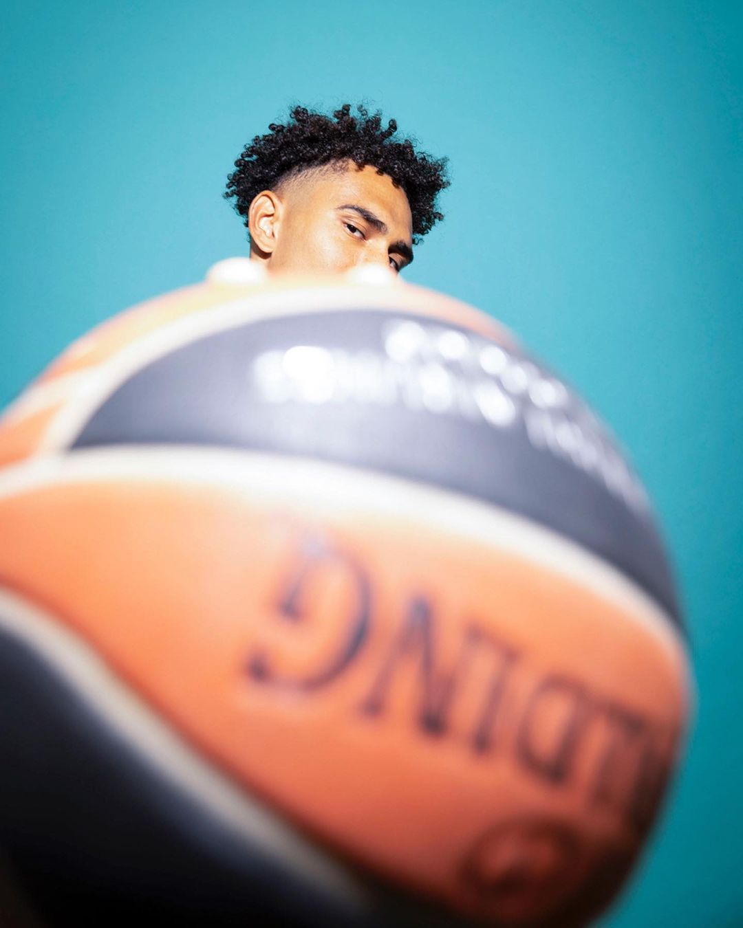 Close-up portrait of a dark-haired man partially obscured by a basketball against a bright teal background, emphasizing sport and portraiture.