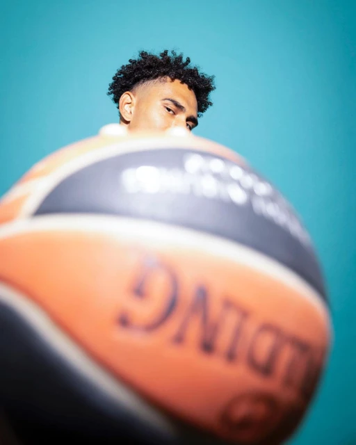 Close-up portrait of a dark-haired man partially obscured by a basketball against a bright teal background, emphasizing sport and portraiture.