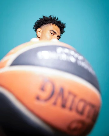 Close-up portrait of a dark-haired man partially obscured by a basketball against a bright teal background, emphasizing sport and portraiture.