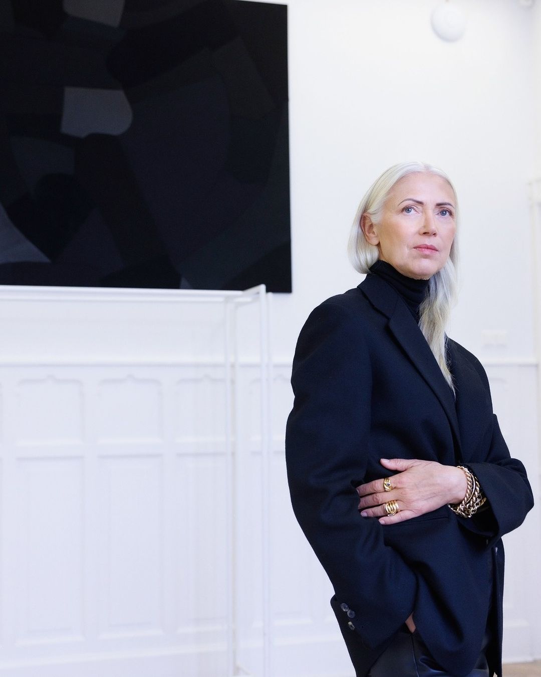 Portrait of a mature adult female with silver hair in a black blazer, gold jewelry, and dark turtleneck against a white wall and artwork.