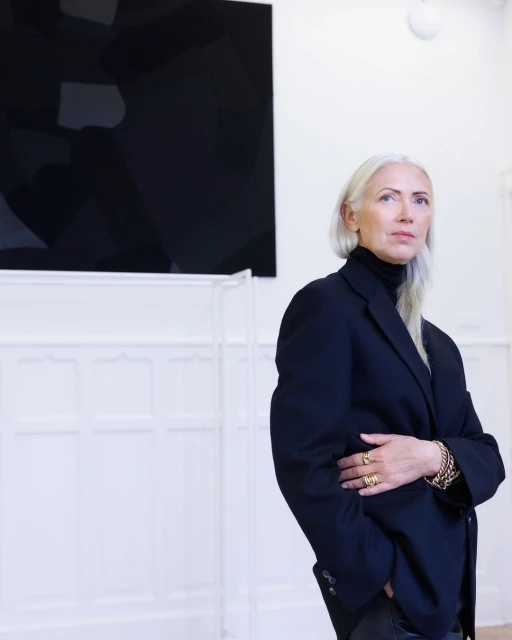Portrait of a mature adult female with silver hair in a black blazer, gold jewelry, and dark turtleneck against a white wall and artwork.