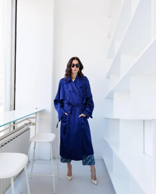 Fashionable woman in a blue trench coat poses indoors near white shelves, wearing sunglasses and heels, showcasing a modern style.