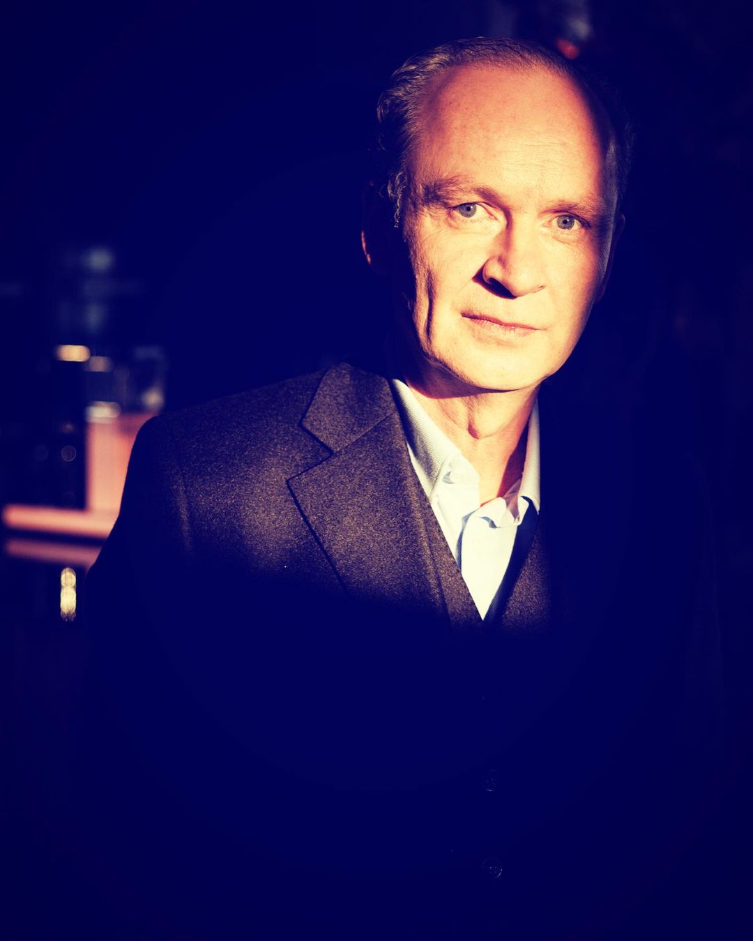 Portrait of a mature adult man wearing a suit jacket and collared shirt, captured in a studio with dramatic lighting.