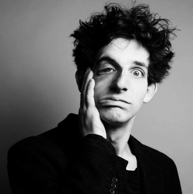 Black and white portrait of a young adult man with dark, messy hair resting his face on his hand, wearing a dark coat against a grey backdrop.