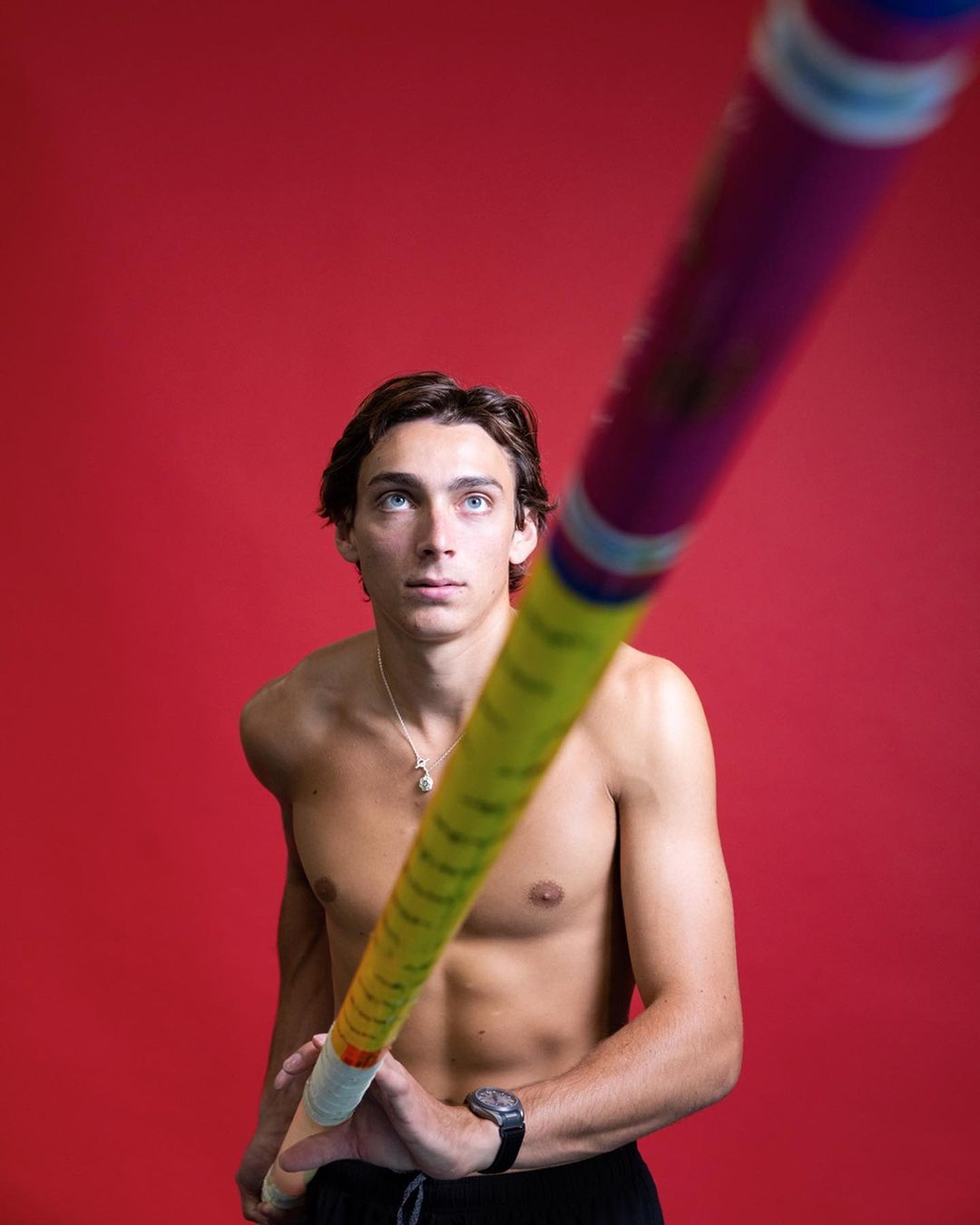 Shirtless pole vaulter in a studio shot with a red background, holding a long yellow pole with pink and blue accents. 