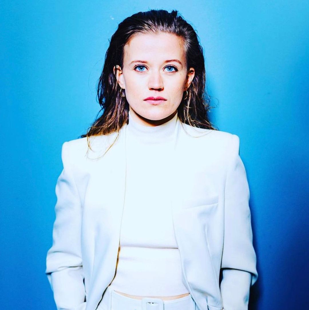 Portrait of a blonde woman with blue eyes, wearing a cream-colored jacket and turtleneck against a bright blue backdrop in a studio setting.