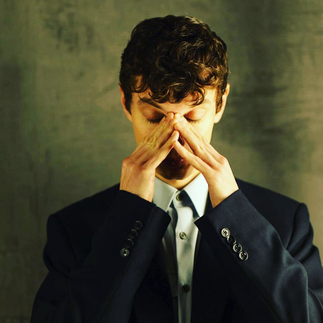Adult male in a suit with curly hair with hands on his forehead in an indoor setting with a textured grey background, portrait.