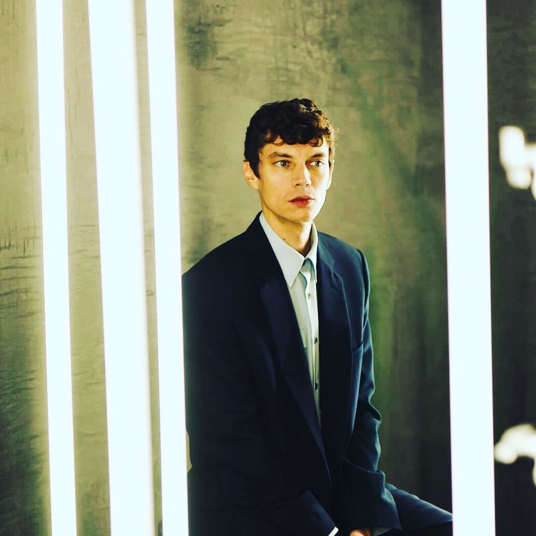 Portrait of a young adult man in a dark blue suit and light blue shirt, posing in front of a pale grey wall with vertical lights.