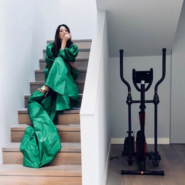 A woman sits on wooden stairs in a long, bright-green satin gown with an elliptical exercise machine in the background indoors.