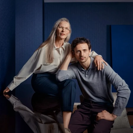 Portrait of an older woman with long grey hair and glasses sitting next to a younger man wearing a grey sweater in a blue interior.