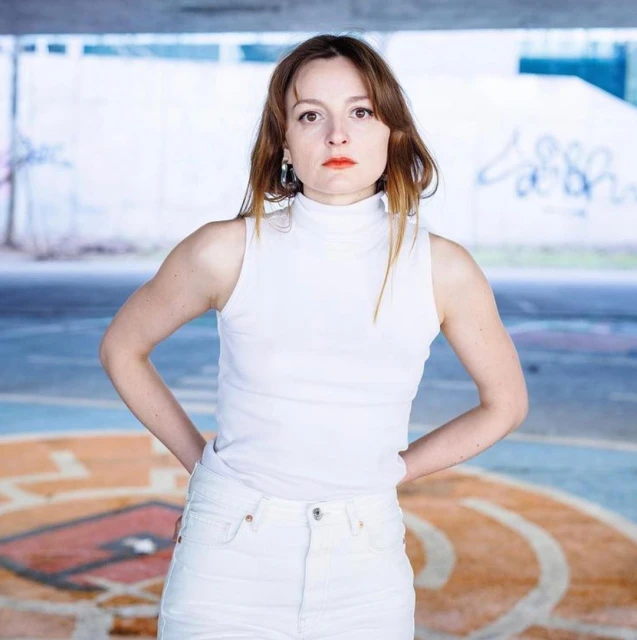 Portrait of a woman in a white top and jeans, posing confidently outdoors under a concrete structure with colorful graffiti.