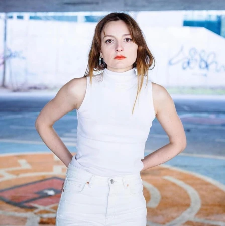 Portrait of a woman in a white top and jeans, posing confidently outdoors under a concrete structure with colorful graffiti.