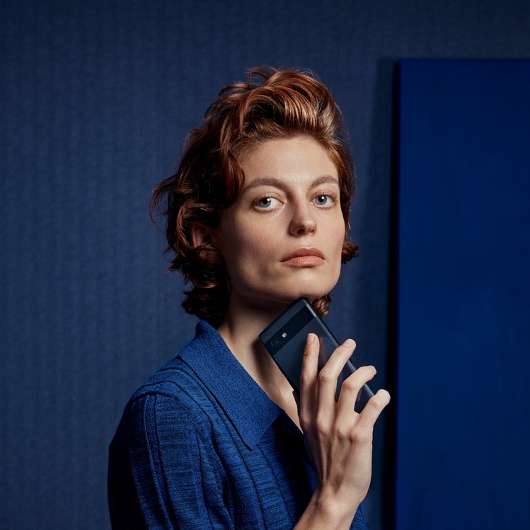 Portrait of a woman with red hair holding a cell phone in front of her chin, wearing a blue textured shirt, set against a blue backdrop.