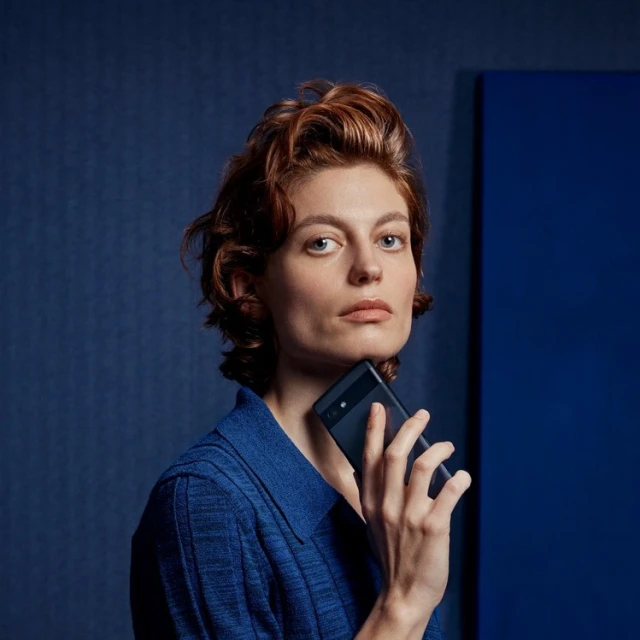 Portrait of a woman with red hair holding a cell phone in front of her chin, wearing a blue textured shirt, set against a blue backdrop.