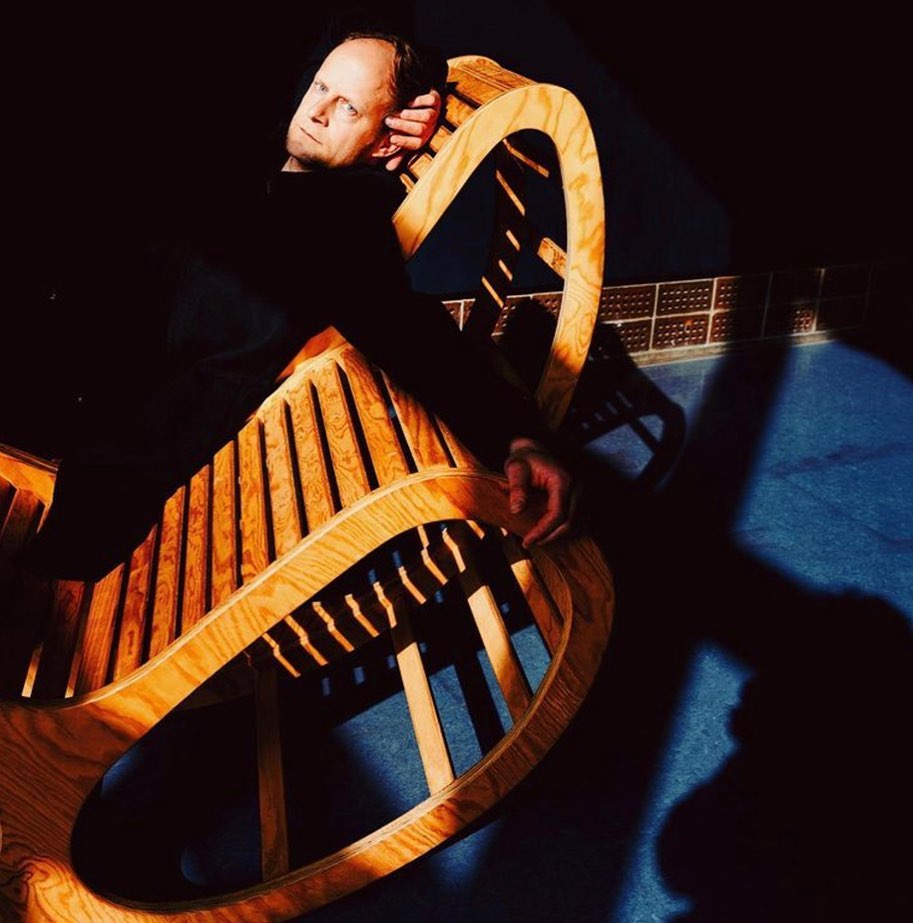 Adult male relaxing on a modern wooden chair in dark lighting with a patterned background, leisure, lifestyle, and creative profession.