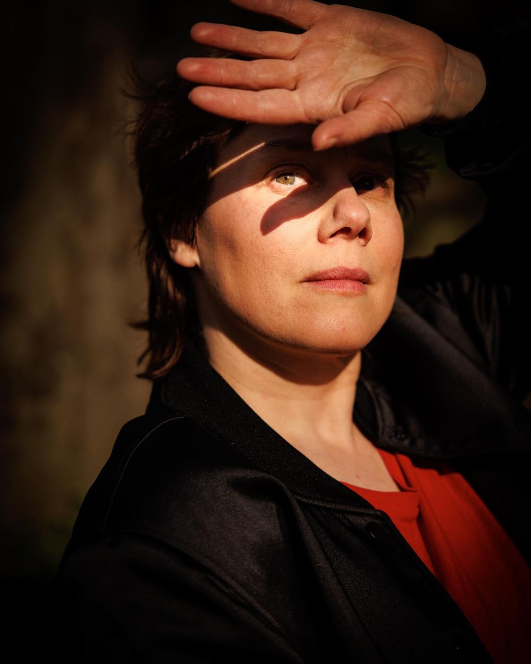 Portrait of an adult woman with her hand shielding her eyes from the sun, wearing a red shirt and black jacket in outdoor environment.