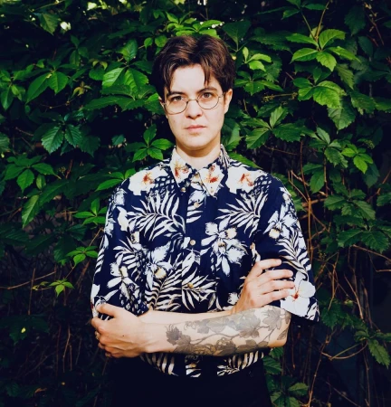 Portrait of a person with short brown hair and glasses wearing a patterned floral shirt with arms crossed against a green leaf background.