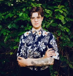 Portrait of a person with short brown hair and glasses wearing a patterned floral shirt with arms crossed against a green leaf background.