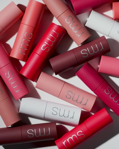 Various lipstick tubes in shades of pink, red, and burgundy are arranged in a scattered pattern on a white surface, brand logo visible.