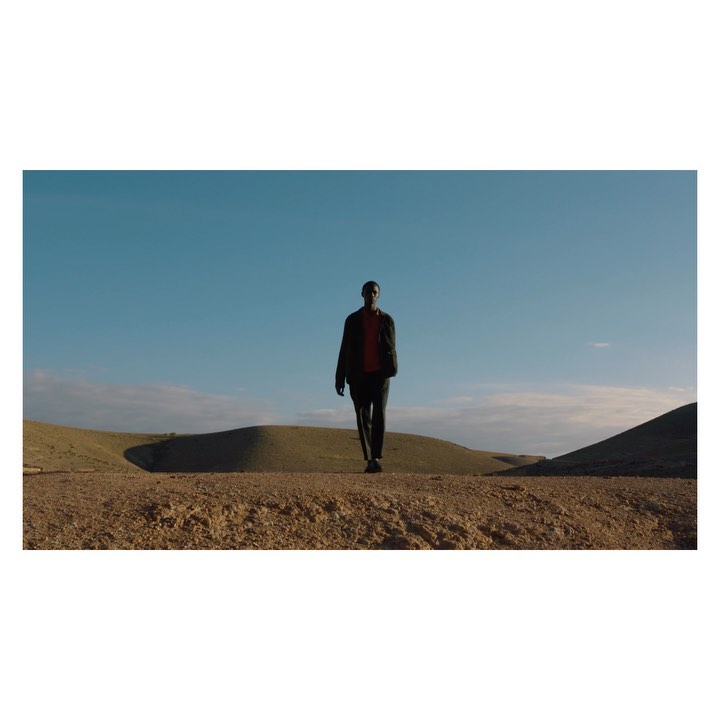 Man walks across arid, hilly terrain under a blue sky with scattered clouds; he is wearing a dark jacket and pants.