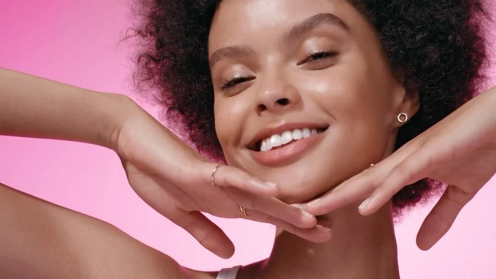 Close-up shot of a smiling adult woman with afro hair and gold rings posing against a pink gradient background in a beauty shot.