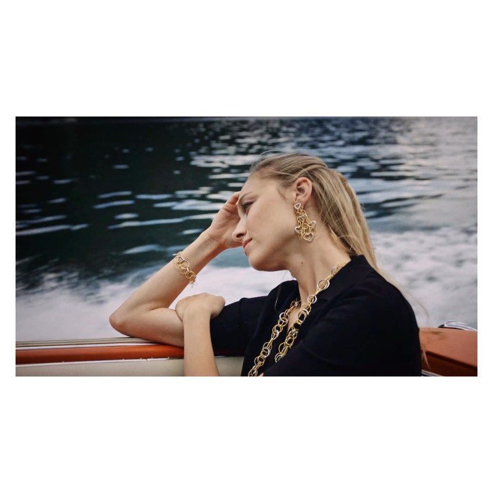 Adult female with blond hair wears gold jewelry on a boat, with dark water in the background. She wears a dark, short-sleeved shirt.