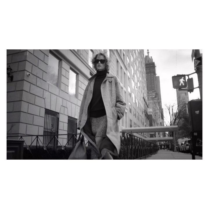Black and white portrait of an adult wearing sunglasses and a coat walking down a city street with buildings and a pedestrian sign.